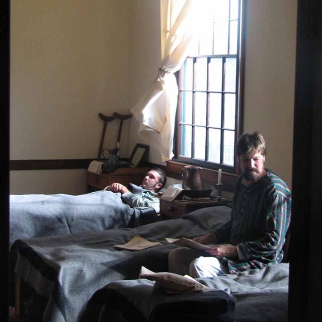 Two men in period dress rest on cots in the post hospital