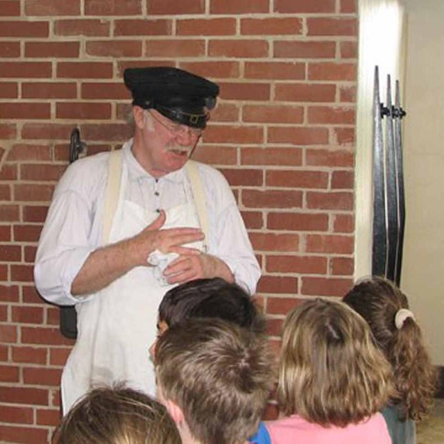A baker in period dress addresses a group of children in front of the bakehouse oven.