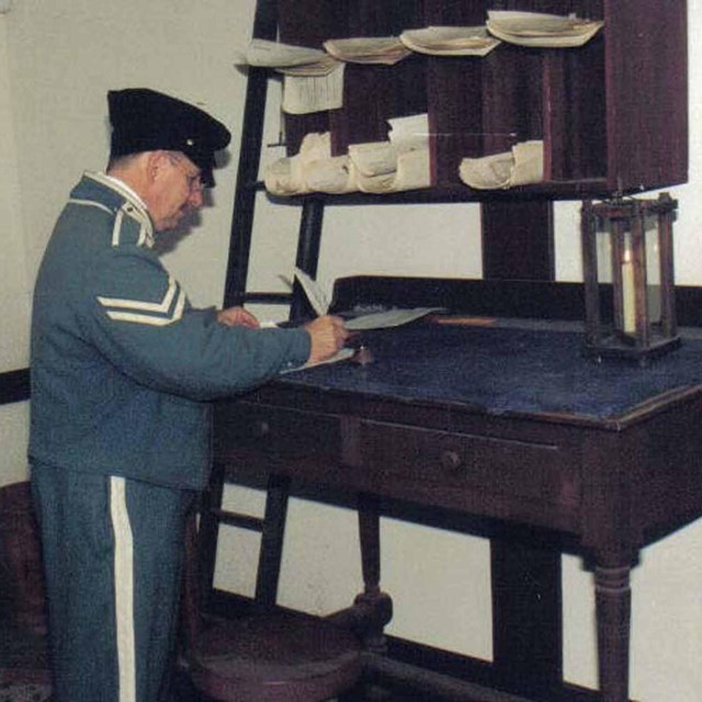 Uniformed quartermaster stands at a desk writing with a quill pen