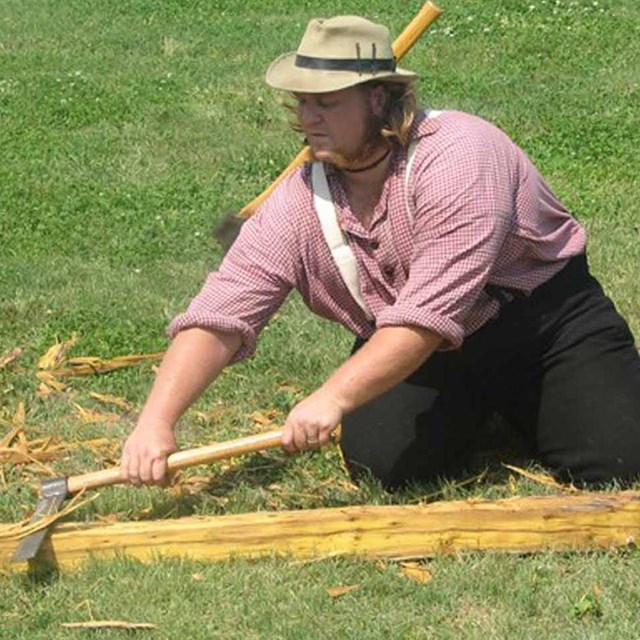 A man in civilian dress cuts a wood beam with an axe