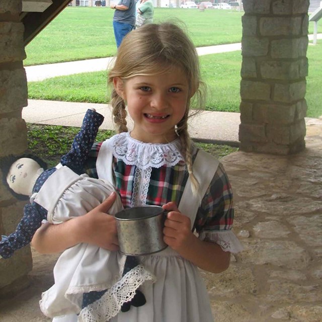 A young girl in period dress holds a rag doll under an awning at Fort Scott