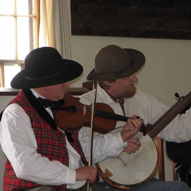 Three men in civilian dress play a fiddle, banjo, and guitar in a building at Fort Scott
