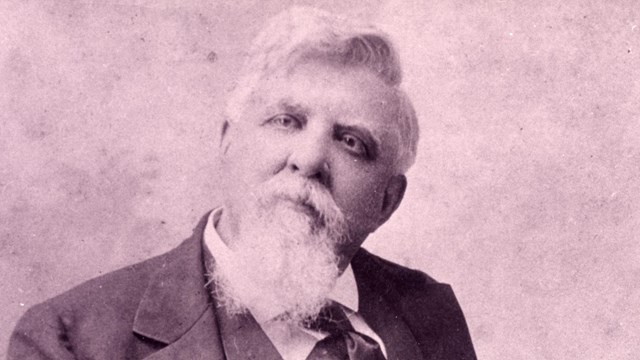 White man with white hair, mustache, and long goatee wearing a dark three piece suit.