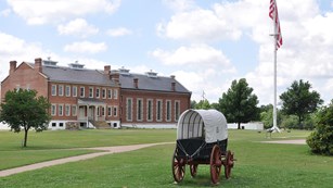 Supply wagon in front of grass parade ground, flagpole and red brick visitor center.