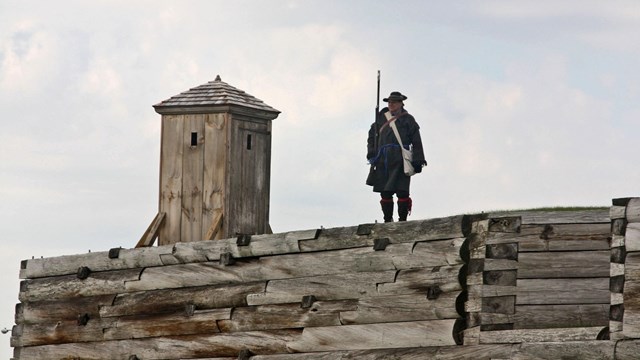 A man in a heavy wool jacket stands with his musket near a sentry box on the fort wall. 