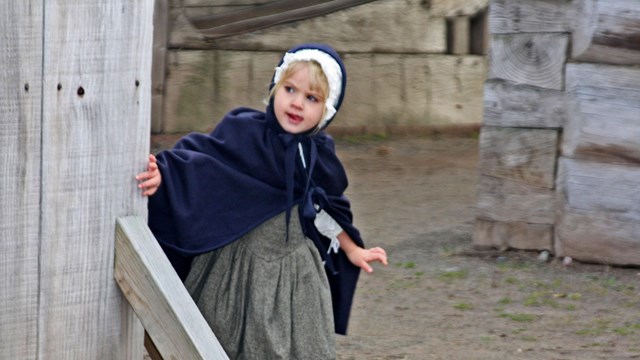 A little girl in 18th C clothing peers at something from behind a sentry box. 