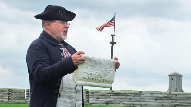 A man in an old-style jacket and tricorn hat reads an old sheet of paper that he holds.