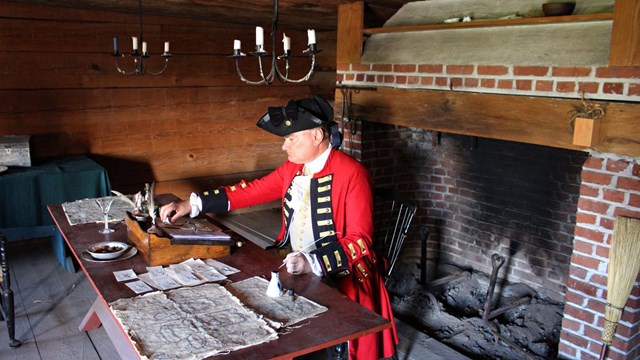 A man in a soldier's uniform sits at a wooden table and writes with a feather pen.