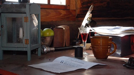 A wooden table with a sheet of paper, a quill pen and ink, and a flute of wine. 