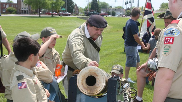 A Continental soldier stands over a brass cannon describing it to scouts. 