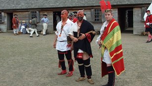 Three Mohawk men stand in the middle of the fort. They are adorned with colorful make-up & clothing.