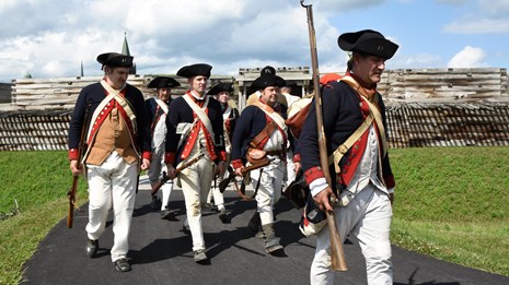 A row of Continental soldiers marches on the path outside the fort wall.