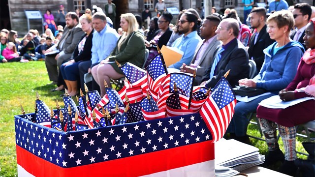A box of flags sits in front of a large, seated crowd. Children sit on the ground nearby. 