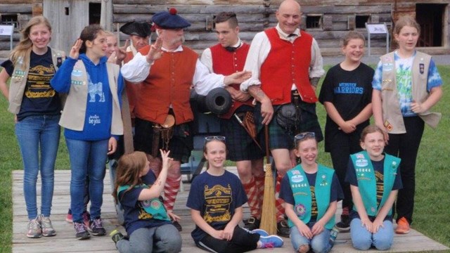 A group of Girl Scouts pose in front of a cannon with people in 18th C clothing. 