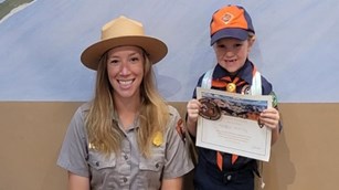 A female blonde ranger poses with a cub scout dressed in an orange and blue uniform.