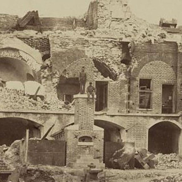 Two soldiers stand and sit on a furnace inside the ruined Fort Sumter.