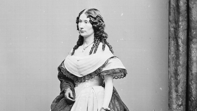 Laura Keene, a white woman in her 30s with dark hair with curls and wearing a large hooped dress.