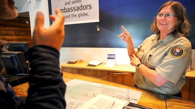 A park volunteer behind a desk communicates with a visitor using sign language.