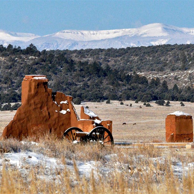 Snow covered mountains behind tree-covered mesa with snow-covered cannon and adobe ruins in front