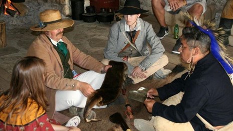 Men and Woman sit in circle on Buffalo Robe examining trade goods and furs.