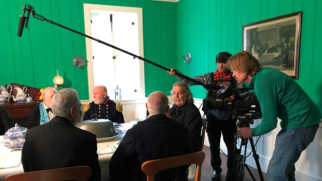 A camera crew films a group of men sitting at the Chief Factor's House table.