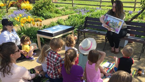 Photo of a preschool-aged children sitting outside listening to an adult read a story.