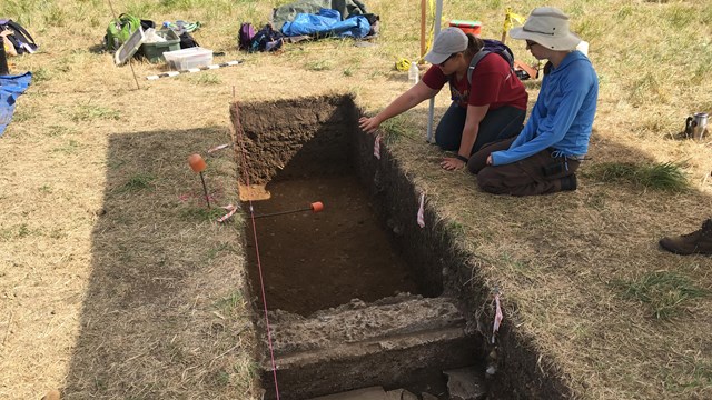 Two archaeologists sit next to an excavation unit.