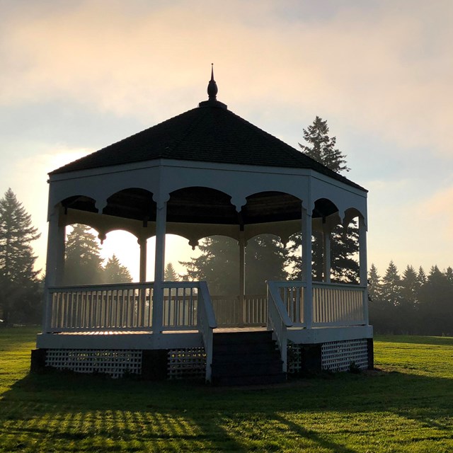A white painted bandstand in a grassy field at sunrise.