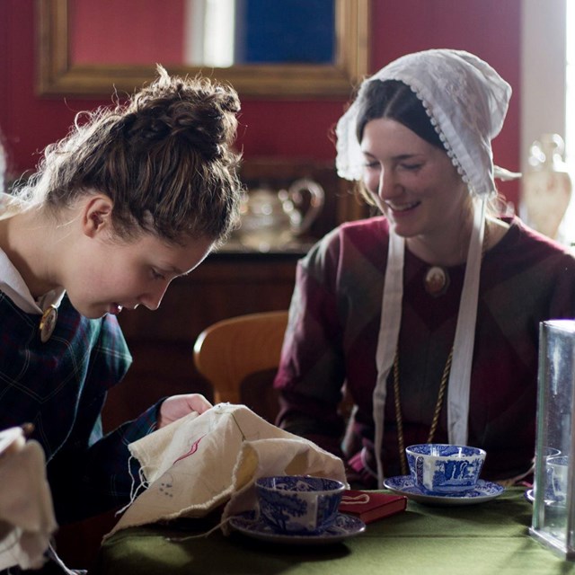 Women doing needlework in 1840s style clothing inside the Chief Factor's House.