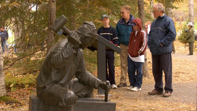 Visitors standing behind a bronze statute of a settler using tools.