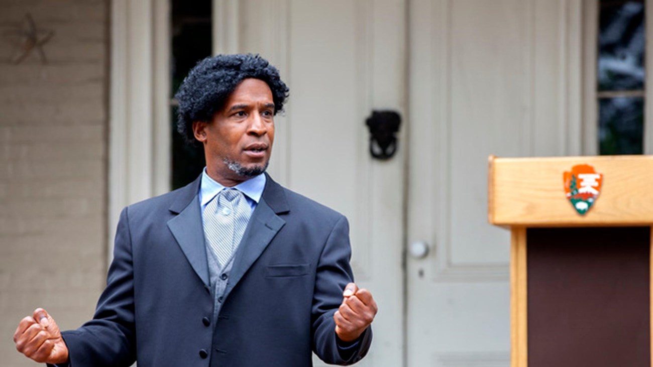 A well-dressed man stands outside of a home, speaking with his hands closed as fists