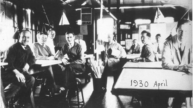 Black and white of group of men seated at tables smiling to camera