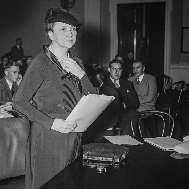 Francis Perkins stands, holding papers while testifying before Congress
