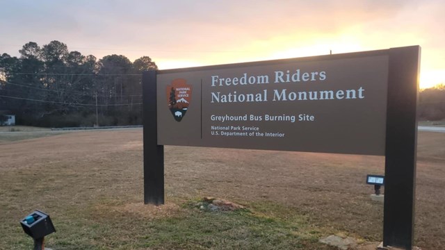 The Freedom Riders National Monument Bus Burning Site sign stands in a field with the sun setting.