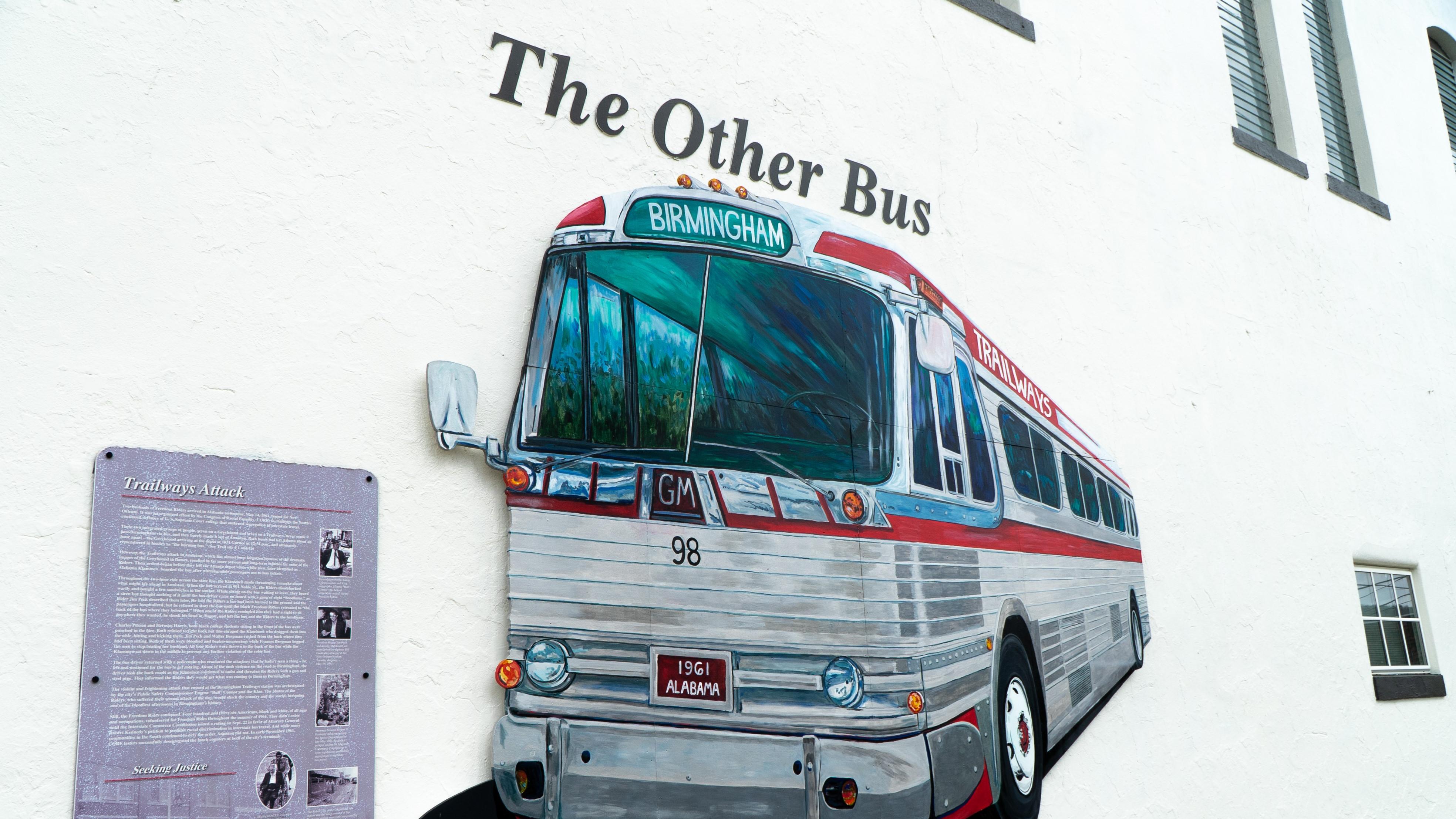 A bus mural labeled "The Other Bus" is painted on a brick wall next to an interpretive panel.
