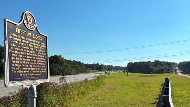 An Alabama historical marker for the Freedom Riders in a grassy field surround by roadways.