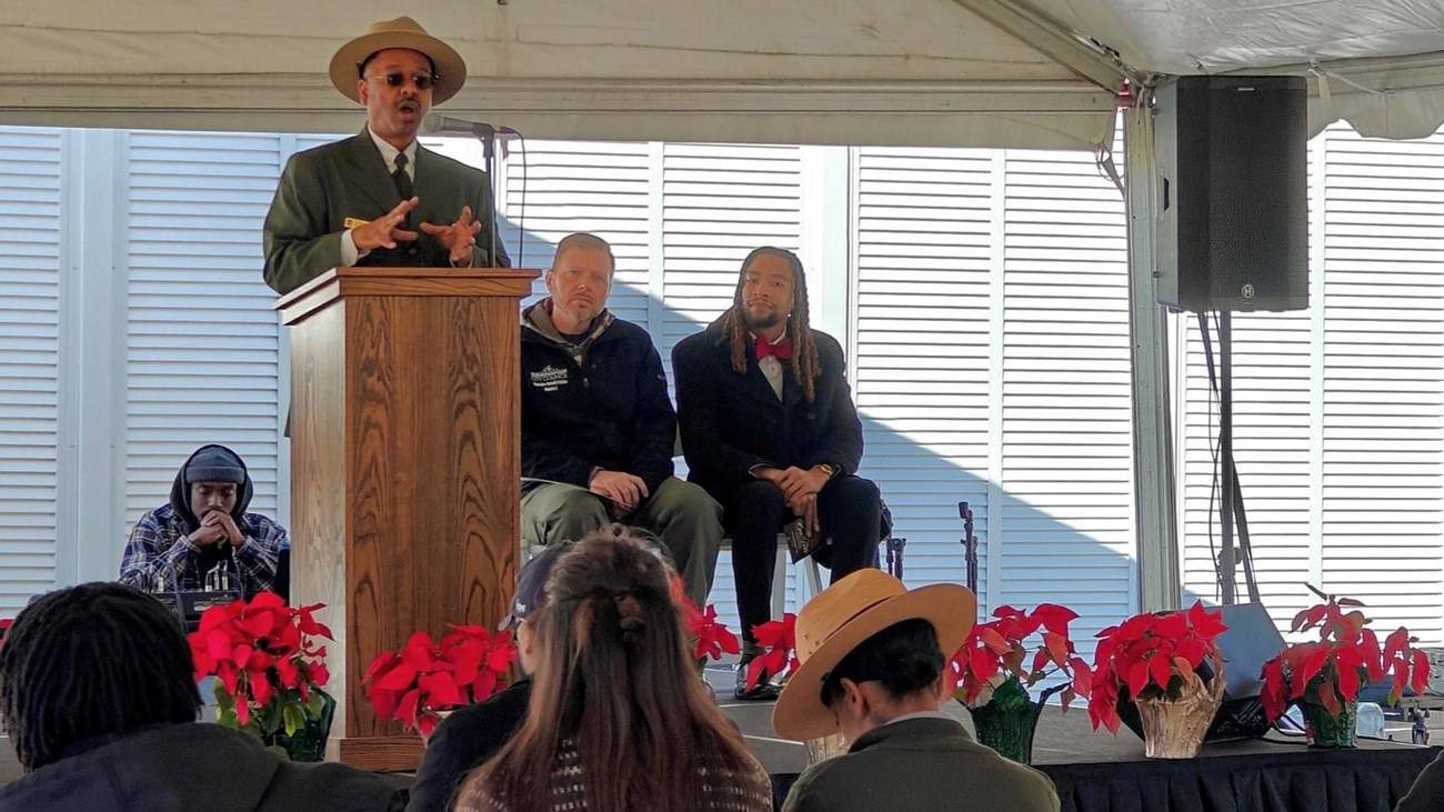 A man speaks on a stage to a group of people. Two people sit behind him. 