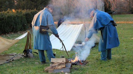 Two living historians dressed as Civil War U.S. soldiers tend to a fire.