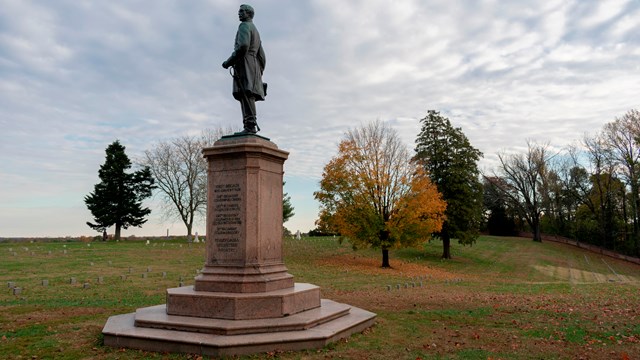 A statue of a man in an open cemetery with fall trees behind.
