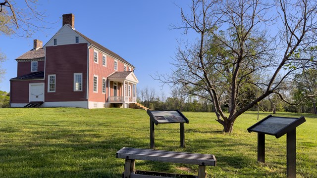 A two-story farm house situated in a field, behind a bench with signs nearby