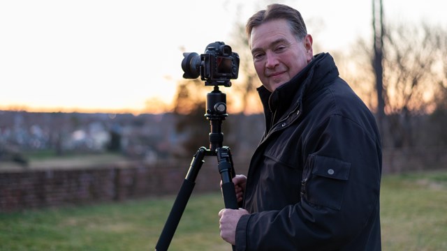 A man standing by a camera, outdoors, at sunset.