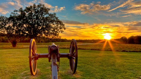 Ruins of house site and cannon with open field and treeline in background while sun sets.