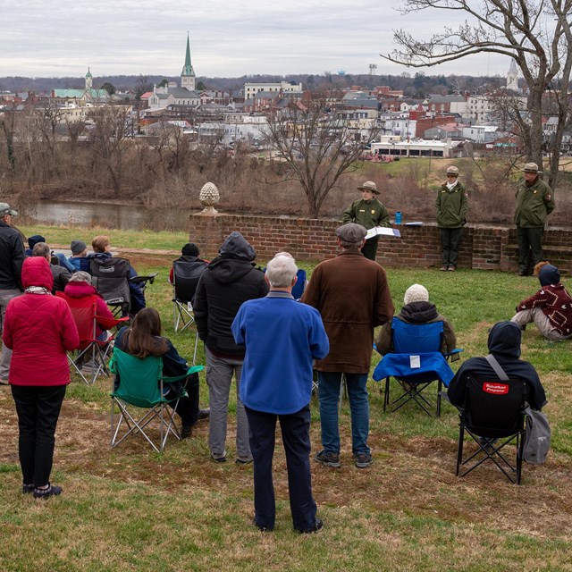 A young female park ranger talks to a group of people by a riverfront with a view of Fredericksburg.