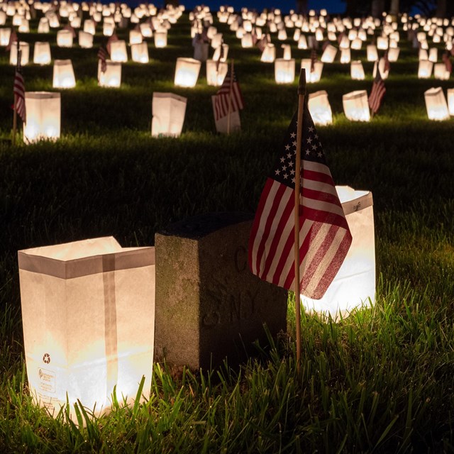 Luminaries surrounding rows and rows of soldier graves decorated with small flags at night 