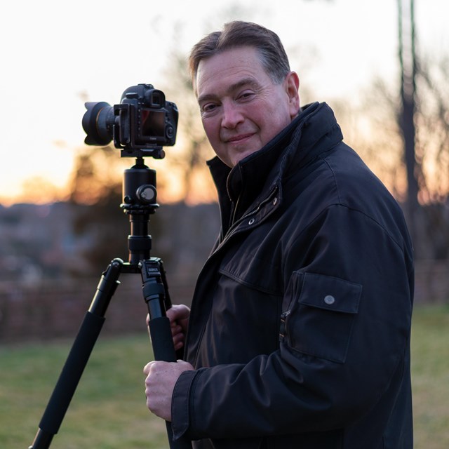 A man standing by a camera, outdoors, at sunset.