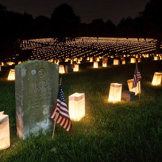 Luminaries surrounding rows and rows of soldier graves decorated with small flags at night 