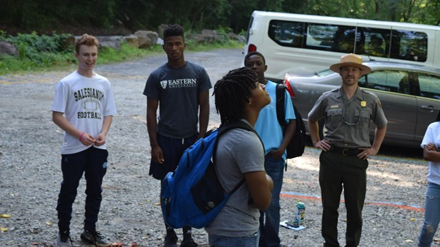 A group of people gather in a gravel parking lot before a program. 