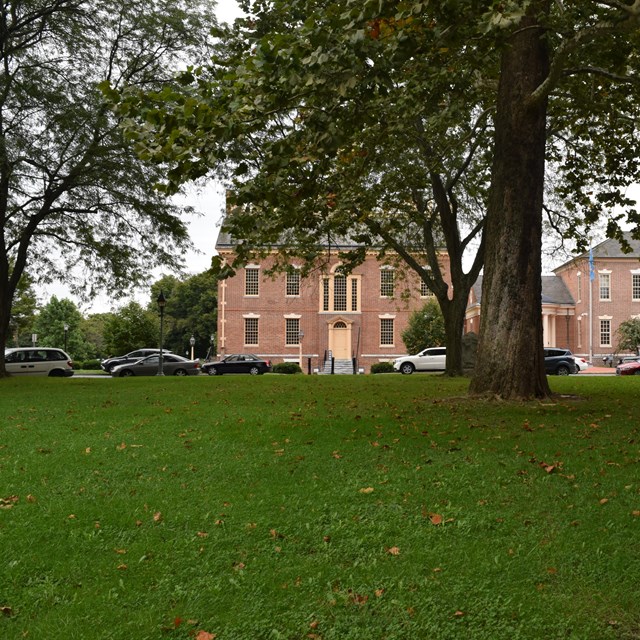 A view looking up from the grass towards a colonial building and large trees. 