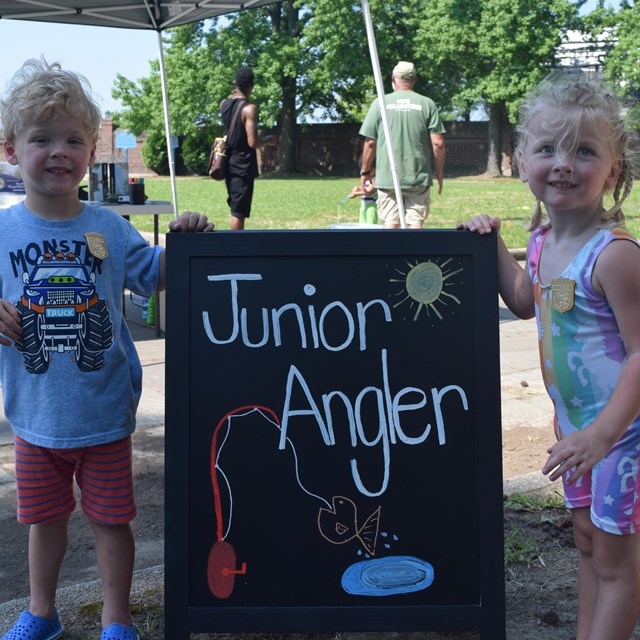 Two young kids pose for a photo in front of a Junior Angler sign. 