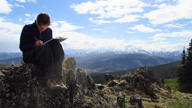 woman sitting on a rock, making notes on a clipboard